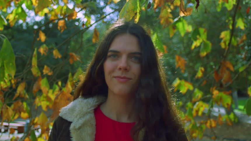 A woman smirks as she stands under a tree that has leaves changing to fall colors
