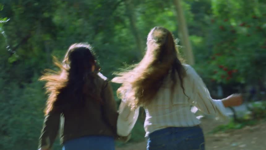 Two women viewed from behind running through a wooded area while holding hands