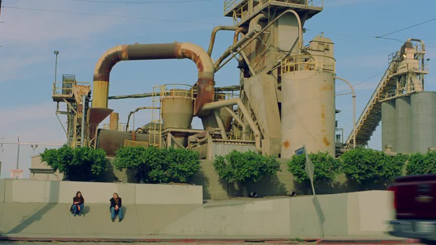 Two women sitting next to a factory facing an urban road with vehicle traffic