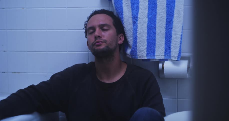 A sad or depressed person is sitting on the bathroom floor taking pills from a prescription bottle