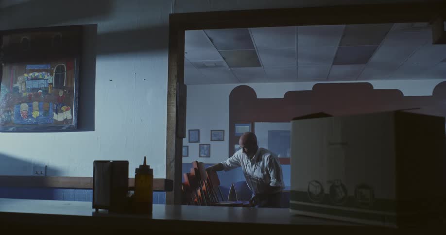 A person is seen through a window inside a dimly lit room stacking chairs on top of tables