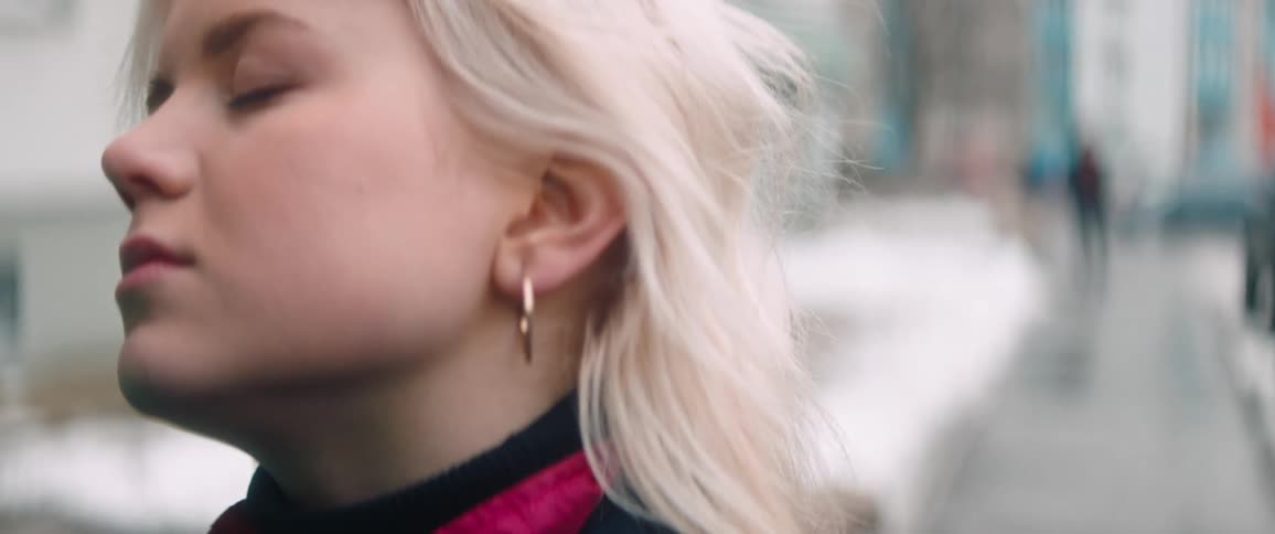 A young woman with blonde hair stands outdoors in front of a city street covered in snow