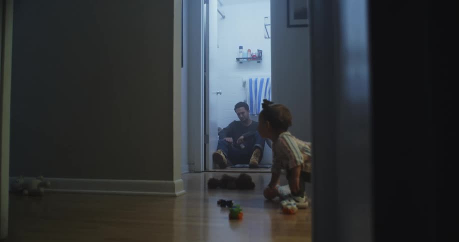 A man is sitting on the bathroom floor as a child picks up a toy in the hall