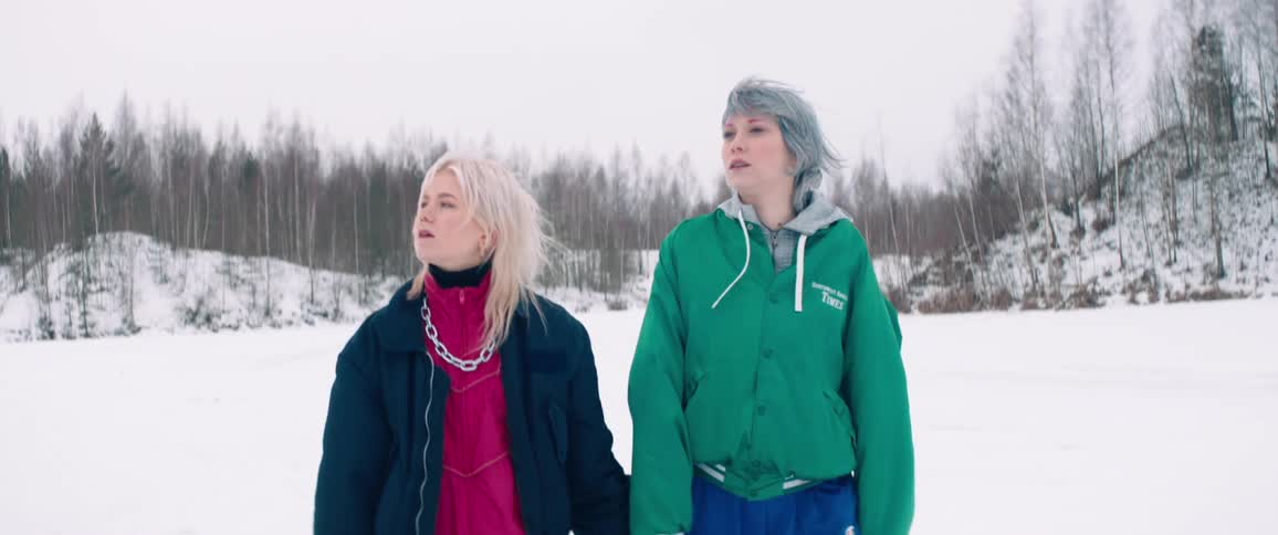 Two women are walking hand in hand in a snowy landscape with a backdrop of leafless trees wearing winter jackets