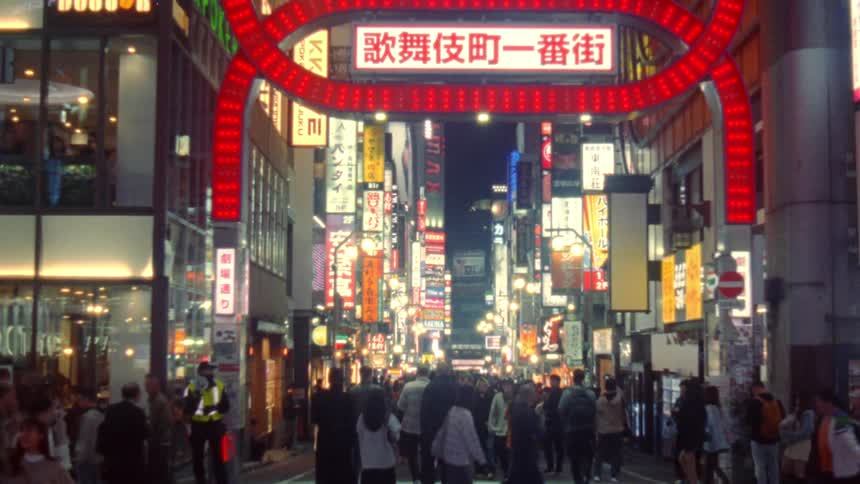 A bustling street at night illuminated by neon lights and filled with people walking past a line of shops and eateries