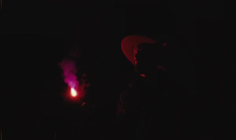 A bearded man wearing a hat holds a red flare in his hand at night