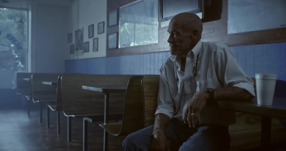An older man is sitting alone at a table in a dimly lit diner with wooden booths