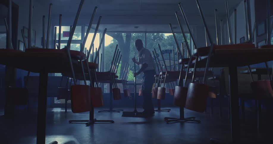 Silhouette of a man cleaning with a broom a room with tables and chairs overturned on top of the tables