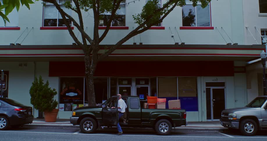 A man is loading boxes and orange chairs onto a green pickup truck parked in front of a store