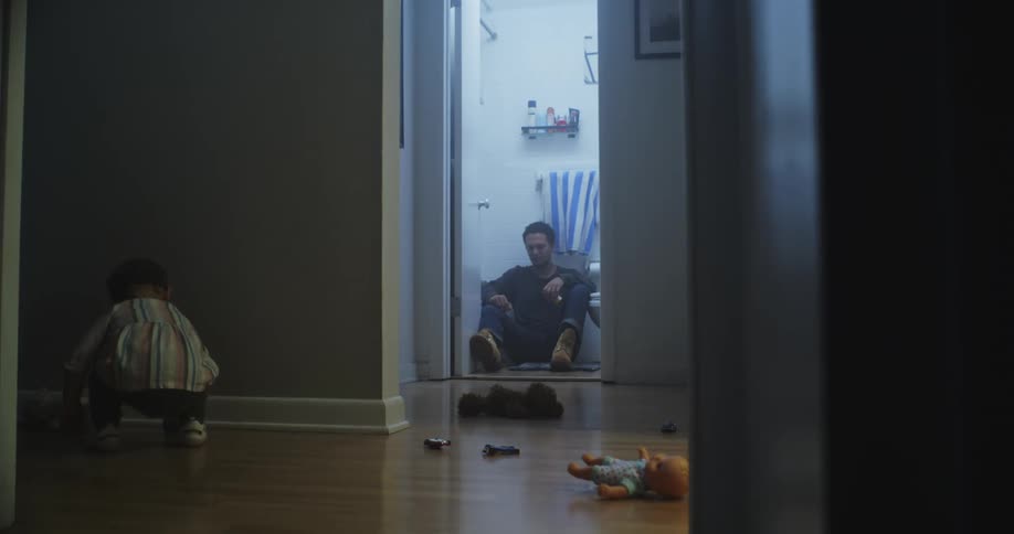 A person is sitting on the floor of a bathroom while a child is crouched on the floor in the hallway surrounded by scattered toys