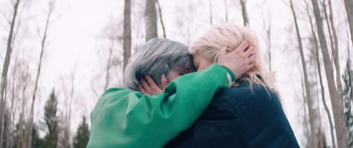 Two women leaning their faces against each other while standing in an outdoor setting with trees