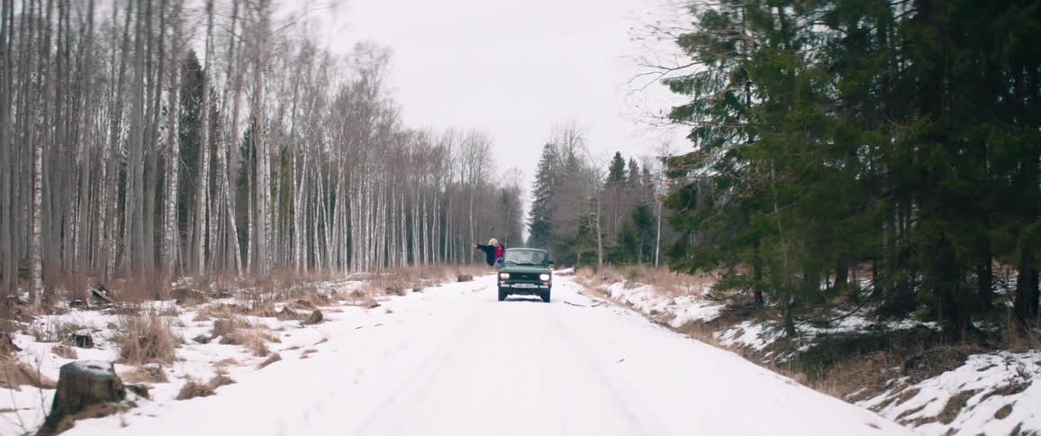 Classic car traveling through a snowy area surrounded by trees and a woman with her arms and body out the window