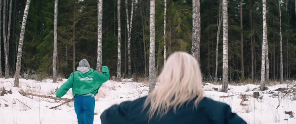 Two women are running through a snowy forest with one in a green jacket leading the way and the other in a dark blue jacket following closely behind