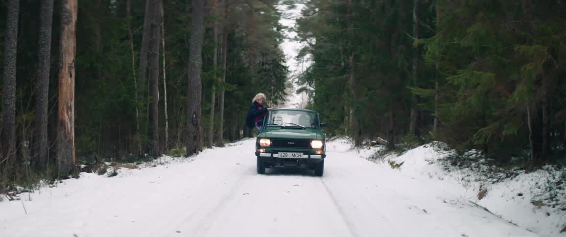 Green car drives along a snowcovered road surrounded by dense forest while a woman excitedly stretches her body out of the window