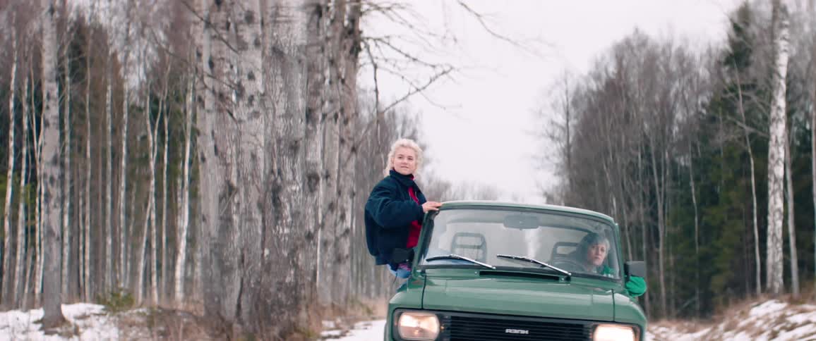 Green classic car driving calmly on a snowy terrain and a woman having fun putting her body out of the window