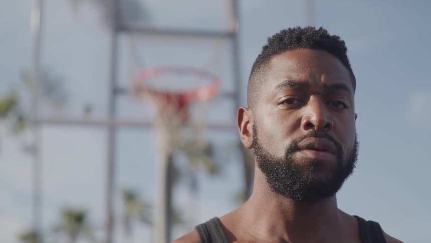 A man with a beard and dark hair is looking forward with a blurred basketball court in the background