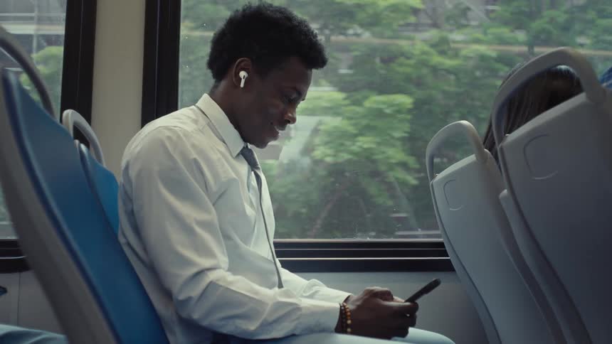 A man wearing a dress shirt and tie sits on public transportation and smiles as he looks at his phone