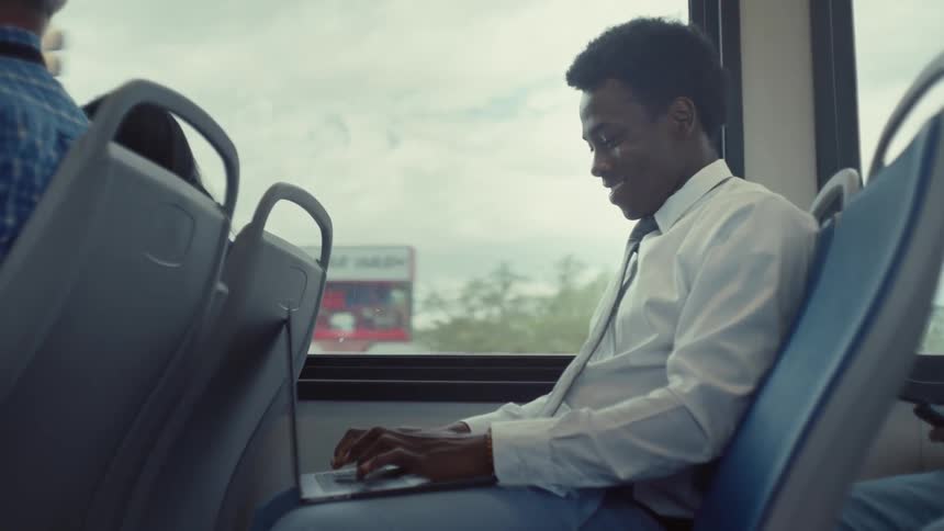 A happy man in a suit types on his laptop while traveling on the bus