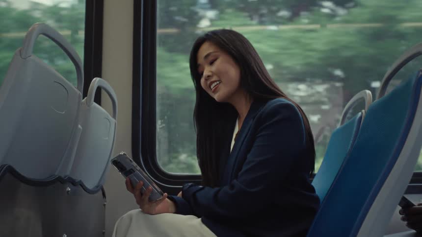 A woman sits on a bus while smiling and holding her smartphone