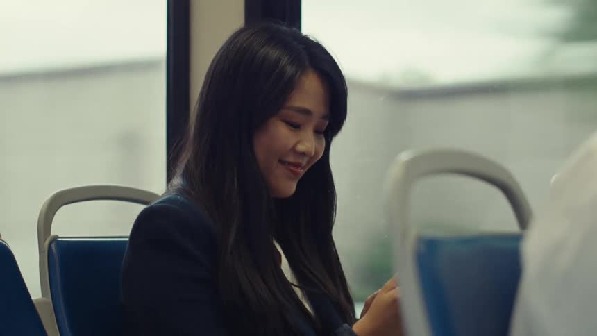 A woman with straight hair and a black blazer answers her phone call while riding the bus