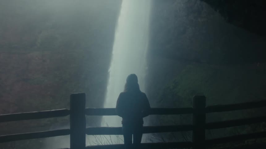 Woman stands on a walkway near an overhang while watching a waterfall