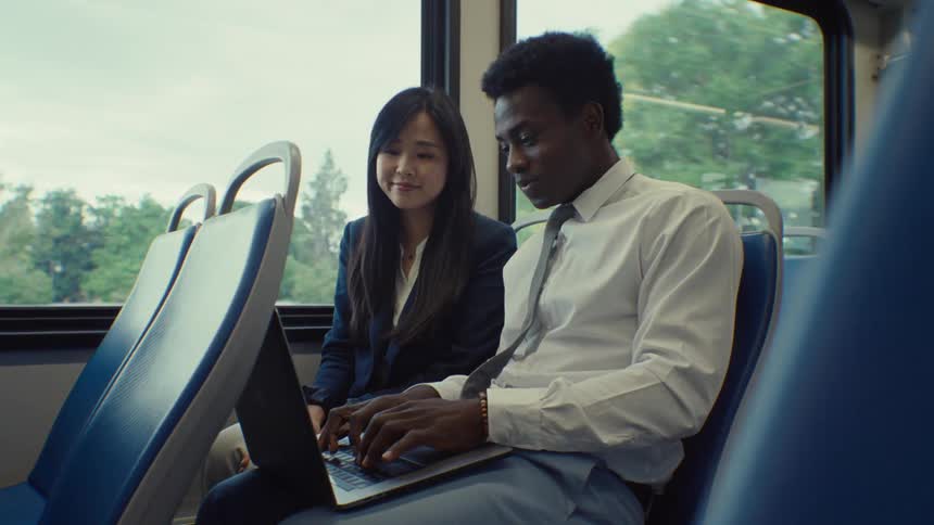 A man and woman ride a train as they talk and he looks up things on the computer
