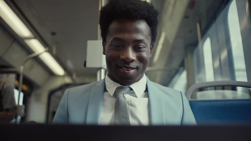 A man wearing a light blue suit and grey tie smiles as he looks at something while riding a train
