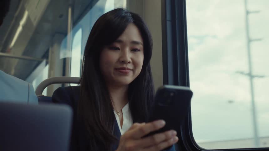 A woman with long dark hair is sitting by a train window looking at her phone