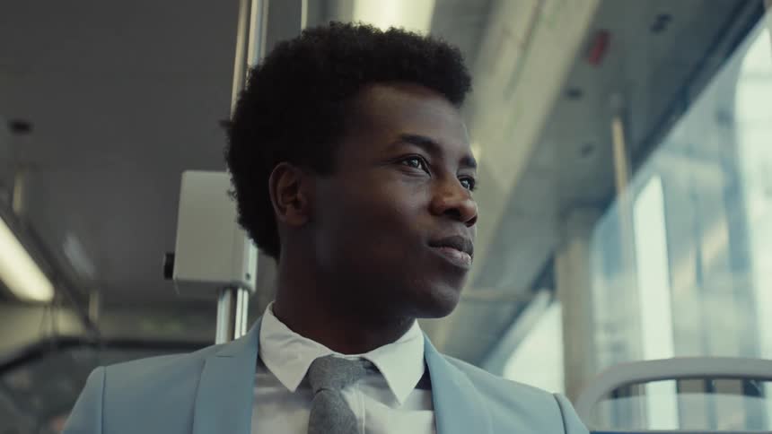 A man in a light blue suit travels on the train gazing out the window with a smile