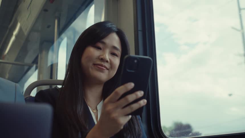 A woman with long dark hair is sitting by a train window looking at her phone