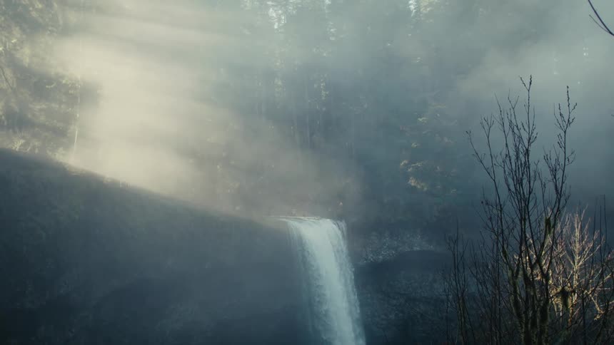 Water falls over the edge of a cliff in a hazy forest