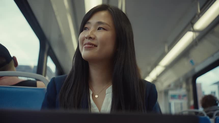 A woman with long dark hair is sitting on a train simling while working on a laptop