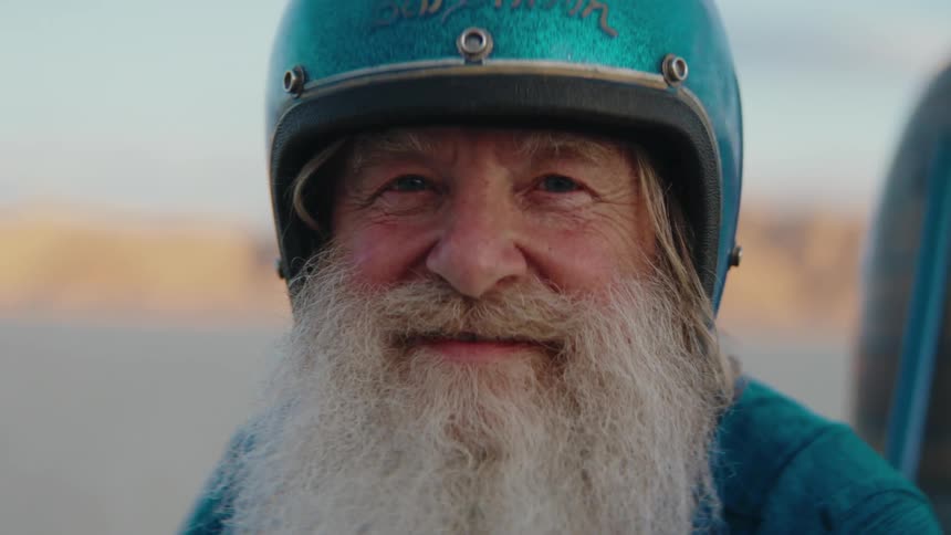 An elderly person with a long white beard wearing a blue helmet and blue clothing stands outdoors with a blurred background