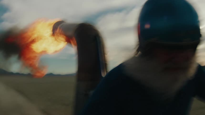 A bearded man wearing a helmet with his beard flowing in the wind drives a jetpowered gokart through the desert