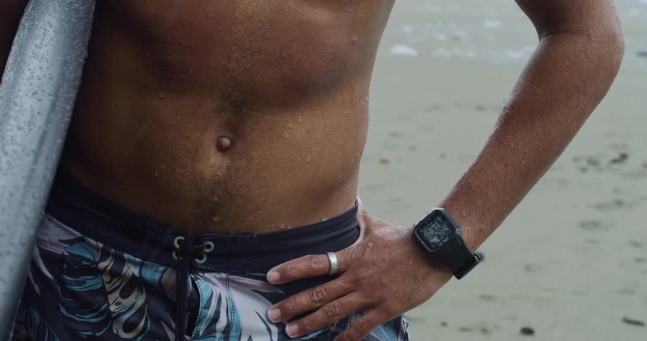 A person is standing on a sandy beach wearing patterned swim trunks and a digital watch with one hand resting on the hip and the other holding a surfboard