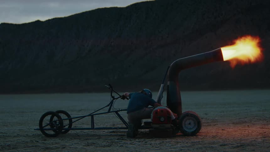 A man in a helmet operating a makeshift vehicle with a mounted flame throwing apparatus set against a dramatic desert landscape during sunset