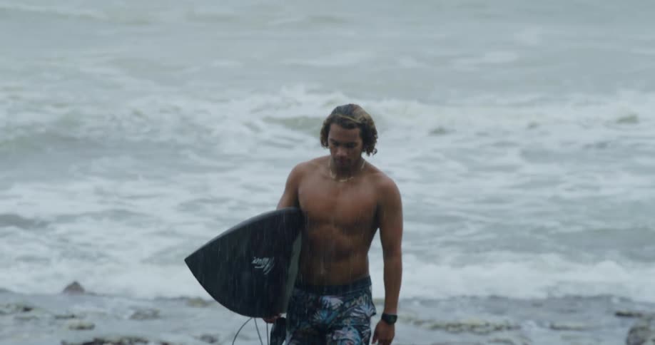 A surfer looks down as he walks away from the water with his surfboard
