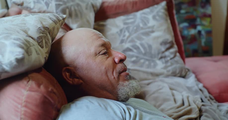 A man is laying against some pillows on a couch and turns to talk