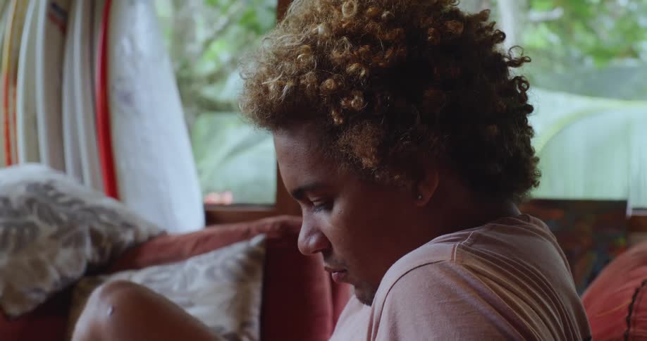 A person with curly hair sits on a couch indoors with surfboards and a lush green outdoor view visible through the window behind them