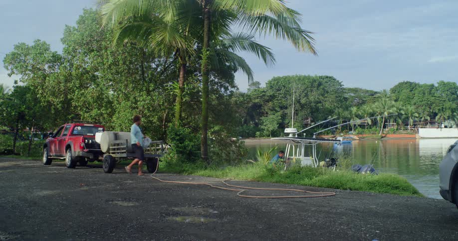 A person on a paved path next to a red pickup truck with a large container on a trailer by a tropical waterfront