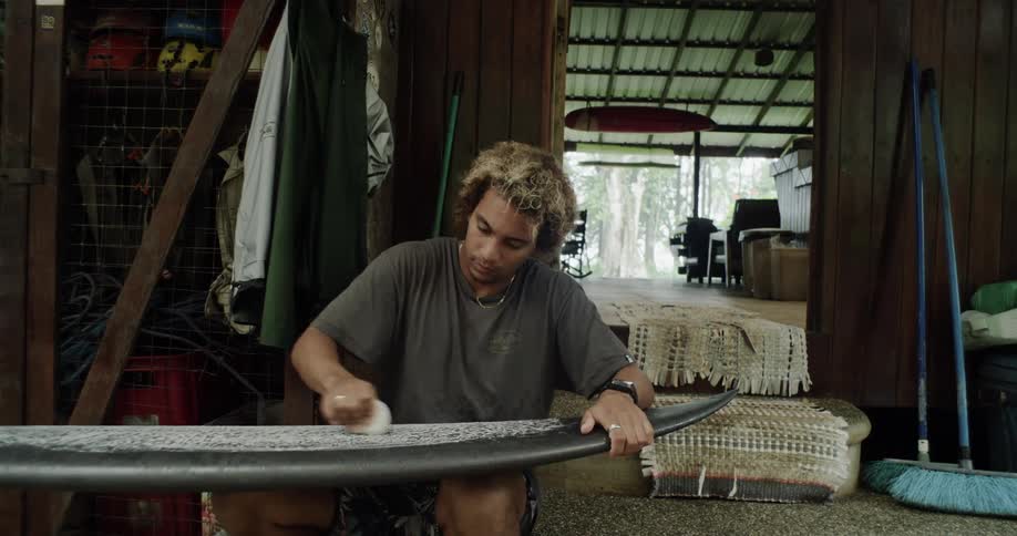 A person is waxing a surfboard in a rustic openair workshop with various tools and equipment in the background