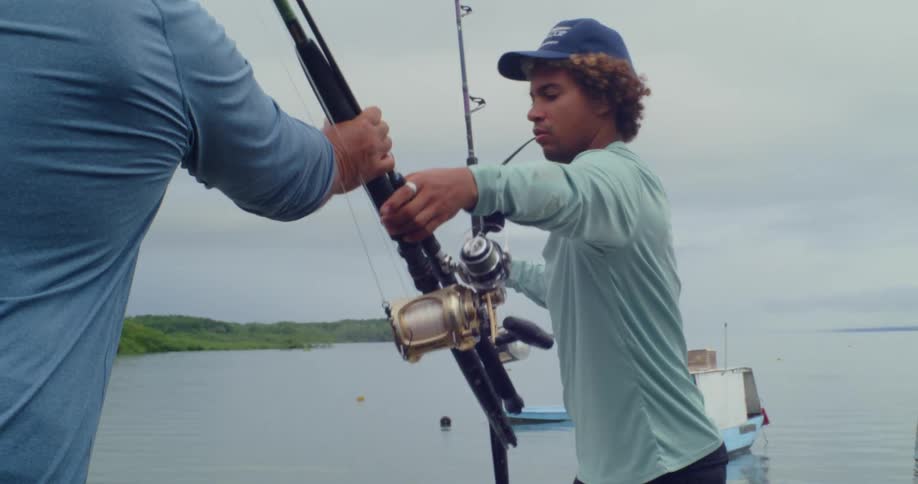 An older man pulls fishing rods out of his pickup truck and hands them to a younger man standing near a lake