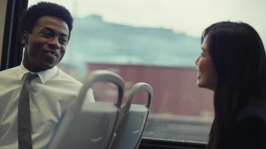 Two people sit on a train conversing with a cityscape visible through the window behind them
