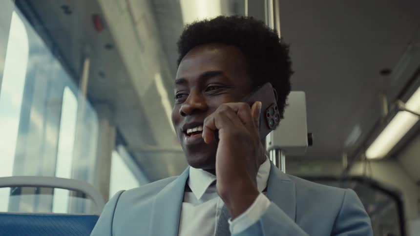A person wearing a light blue suit is sitting on a moving train talking on a smartphone