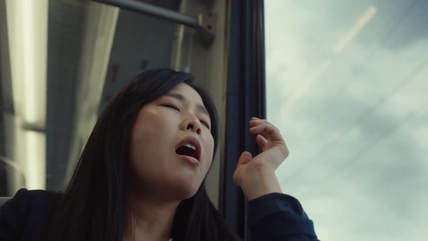 A woman with straight hair and a dark blue blazer jacket travels by train yawning occasionally