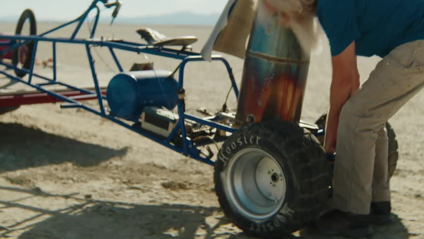 A man works on a dune buggy in a desert area near mountains
