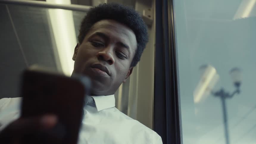 A man wearing a white shirt and grey tie looks out a window then down at his phone while sitting in what appears to be a train car
