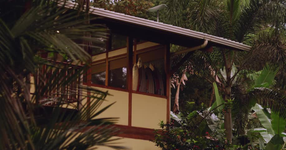 A corner of a twostory house with large windows and a balcony surrounded by lush tropical vegetation