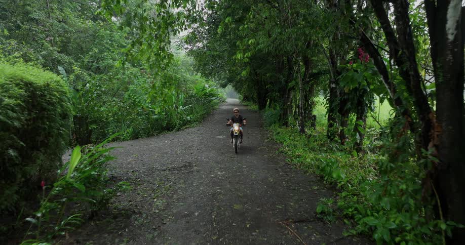 A person is riding a motorcycle down a narrow treelined path surrounded by dense lush vegetation