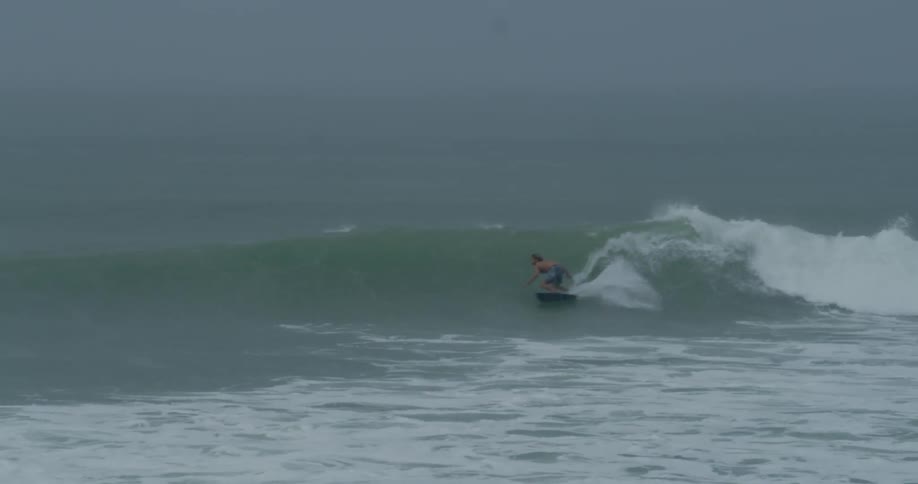 A shirtless man is surfing on a wave formed in a calm sea and doing maneuvers